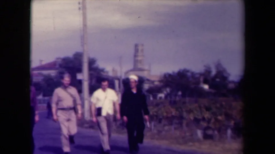 Three adult men, including a likely sailor in a dark uniform and white cap, pass the camera on a rural road in France.