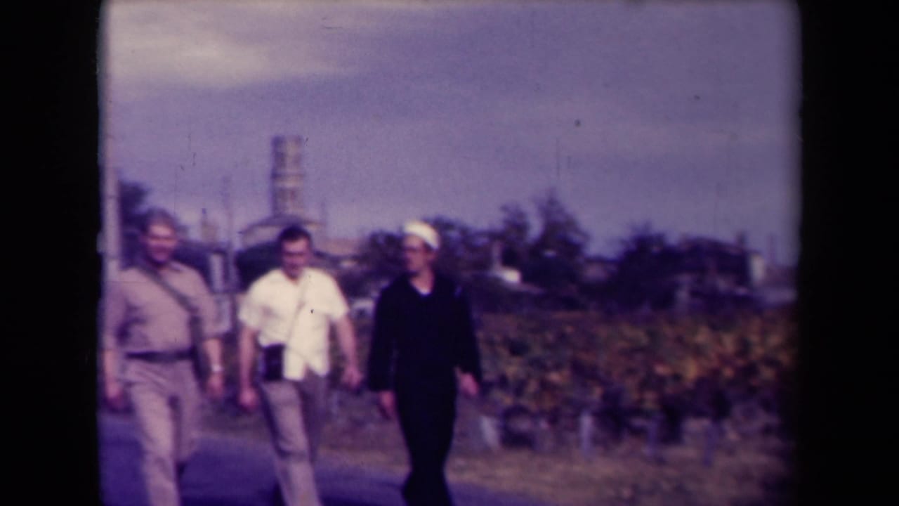 Four adult men walk toward the camera beside a vineyard, with rural structures in the background.