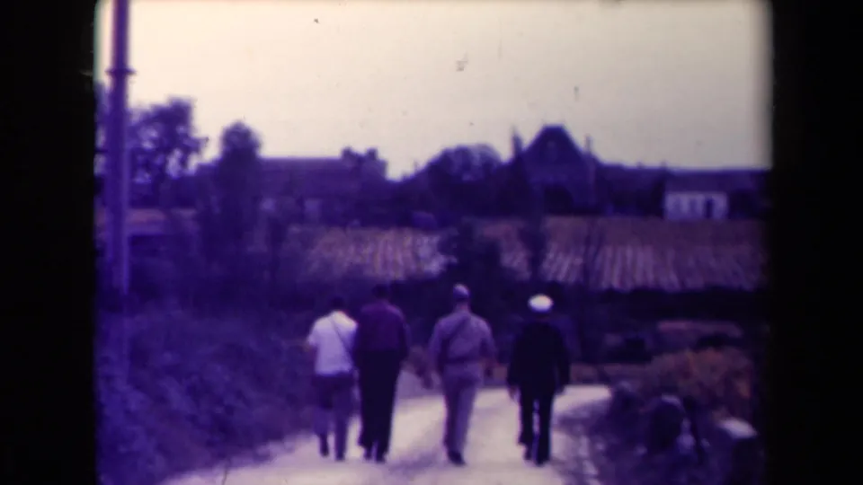 Four adult men are seen from behind on a rural French path, with cultivated rows, a utility pole, trees, houses, and a church tower beyond.