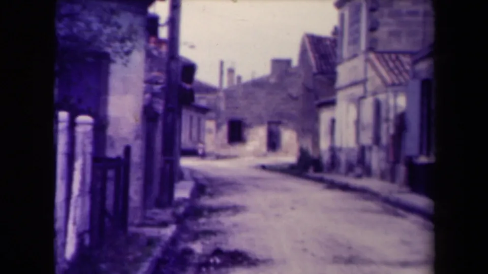 Narrow village street in France, identified as 1945, with old stone buildings and a quiet unpaved road.