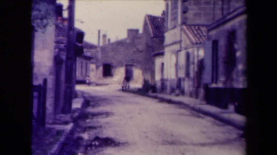 1945 France frame of a lone person carrying something along a quiet street between old buildings.