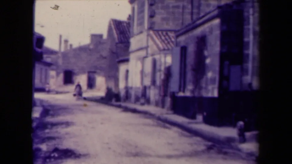 1945 France frame showing a lone distant figure on a narrow street bordered by aged buildings.