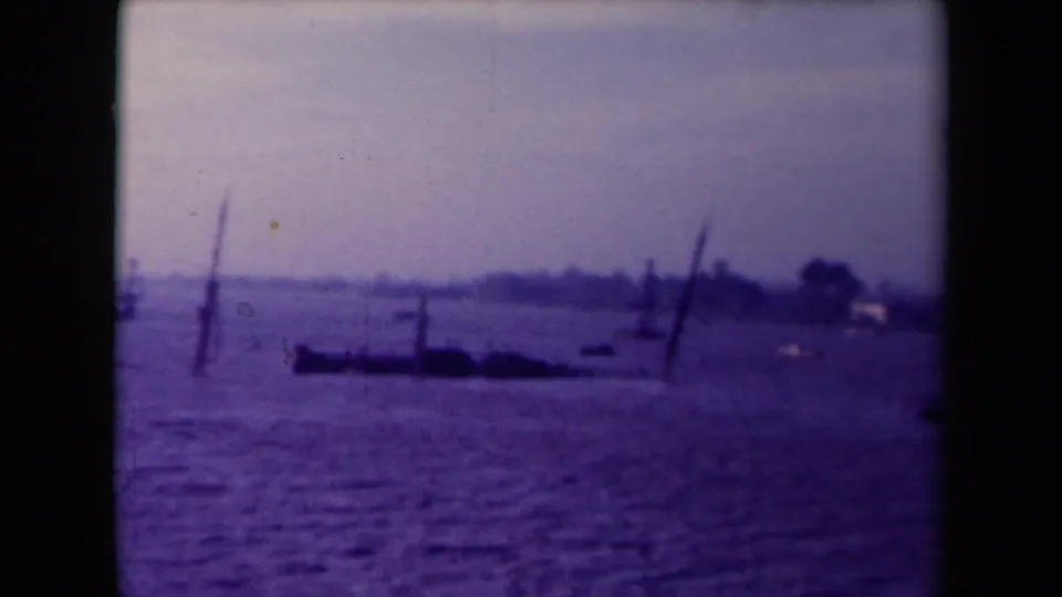 Likely France in the 1940s, with floodwater covering the foreground and thin poles, boats, trees, and pale shoreline buildings beyond.