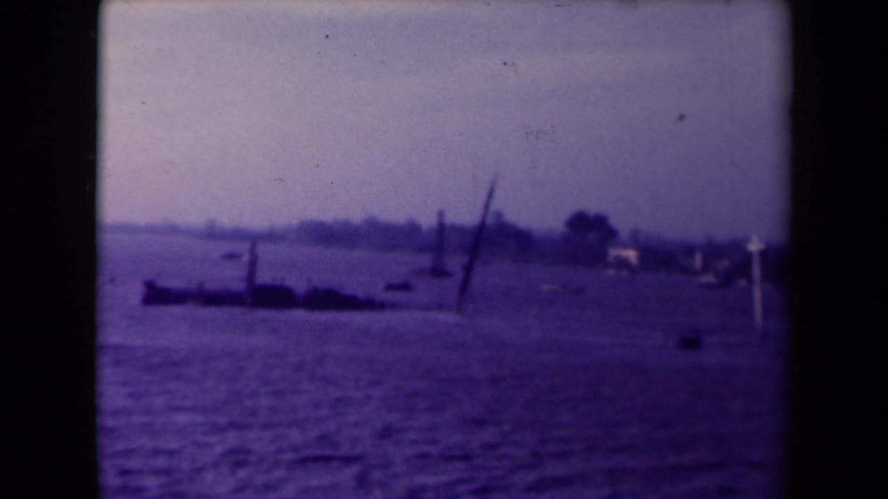 Soft 8mm view of a flooded waterside area, likely France, with a dark low structure, leaning pole, small boats, and distant buildings.