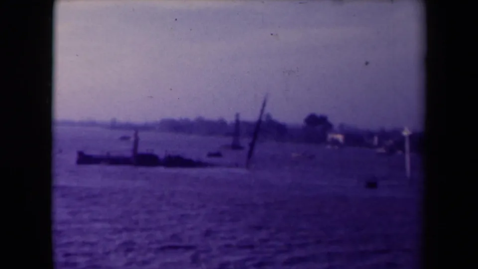 Soft 8mm view of a flooded waterside area, likely France, with a dark low structure, leaning pole, small boats, and distant buildings.