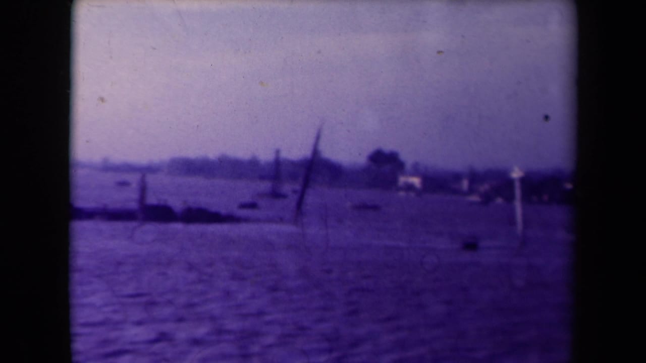 Purple-shifted 1940s 8mm frame, likely France, showing floodwater, scattered posts, small boats, trees, and a pale building on shore.
