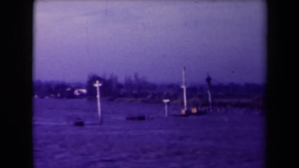1945 France 8mm frame showing white cross markers partly surrounded by floodwater near a low shore.