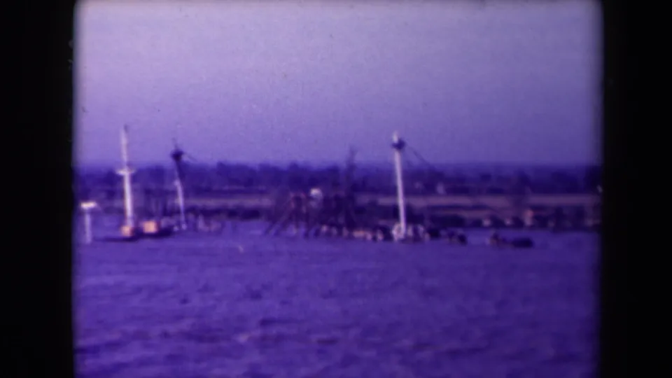1945 France home-movie frame holding on a hazy waterfront with vertical white markers and indistinct boats or barges.