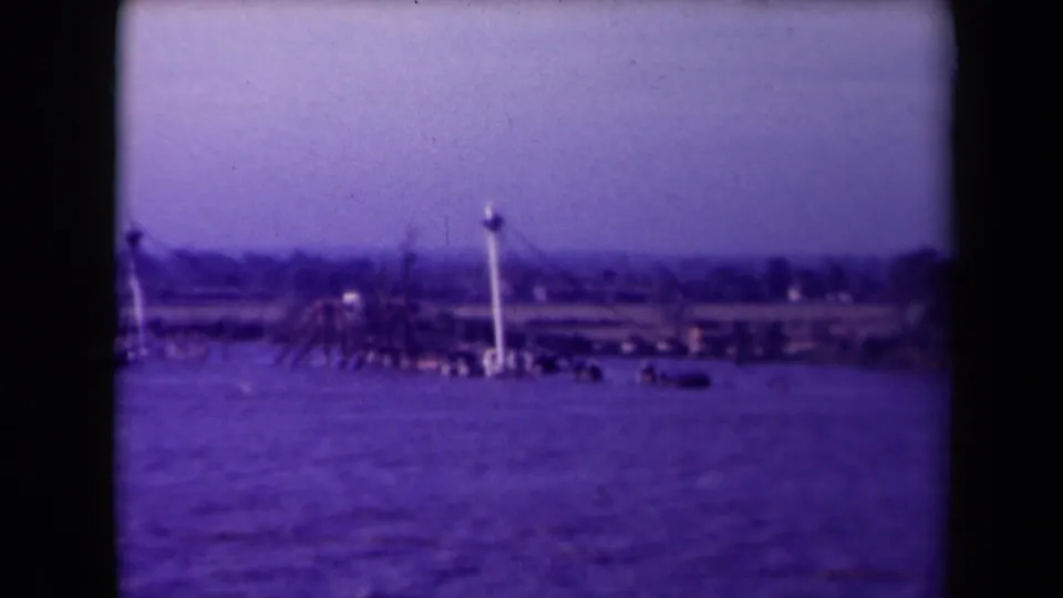 Final 8mm frame of the France coastal view, with rippled water in front of a low shoreline and tall pale waterfront structures.