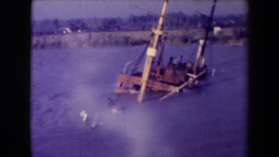 Closer 1945 France frame of the partially submerged wooden boat, its masts tilted and debris floating nearby in the bay.