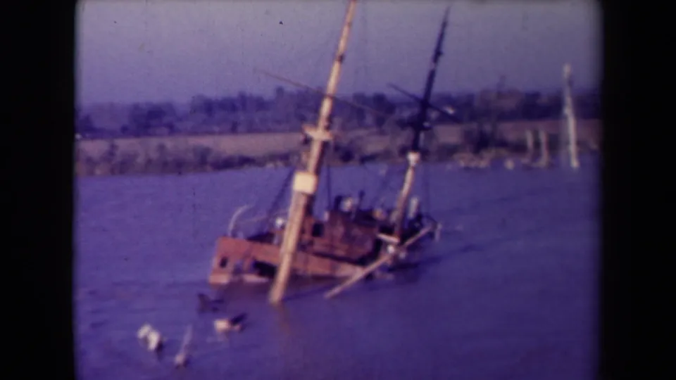 Close 8mm frame from France showing the wrecked wooden boat listing in the bay, with debris and a distant shoreline visible.