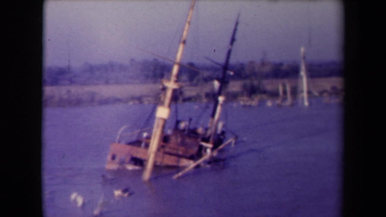 A red and white vessel tilts low in calm water with leaning masts and a distant shoreline behind it.