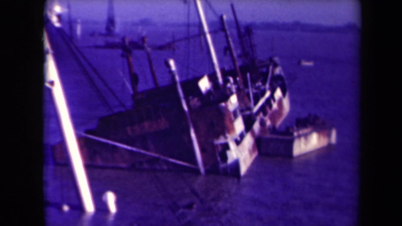 Large tilted shipwreck partly submerged, with masts and rigging over calm water.