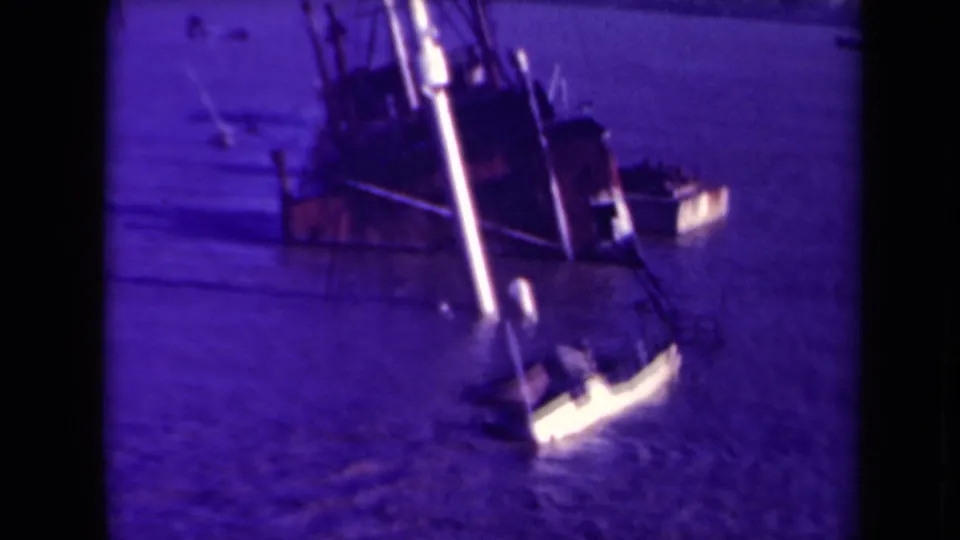 Purple-cast 8mm frame showing the wrecked ship, rigging, water, and a small light-colored boat in France.