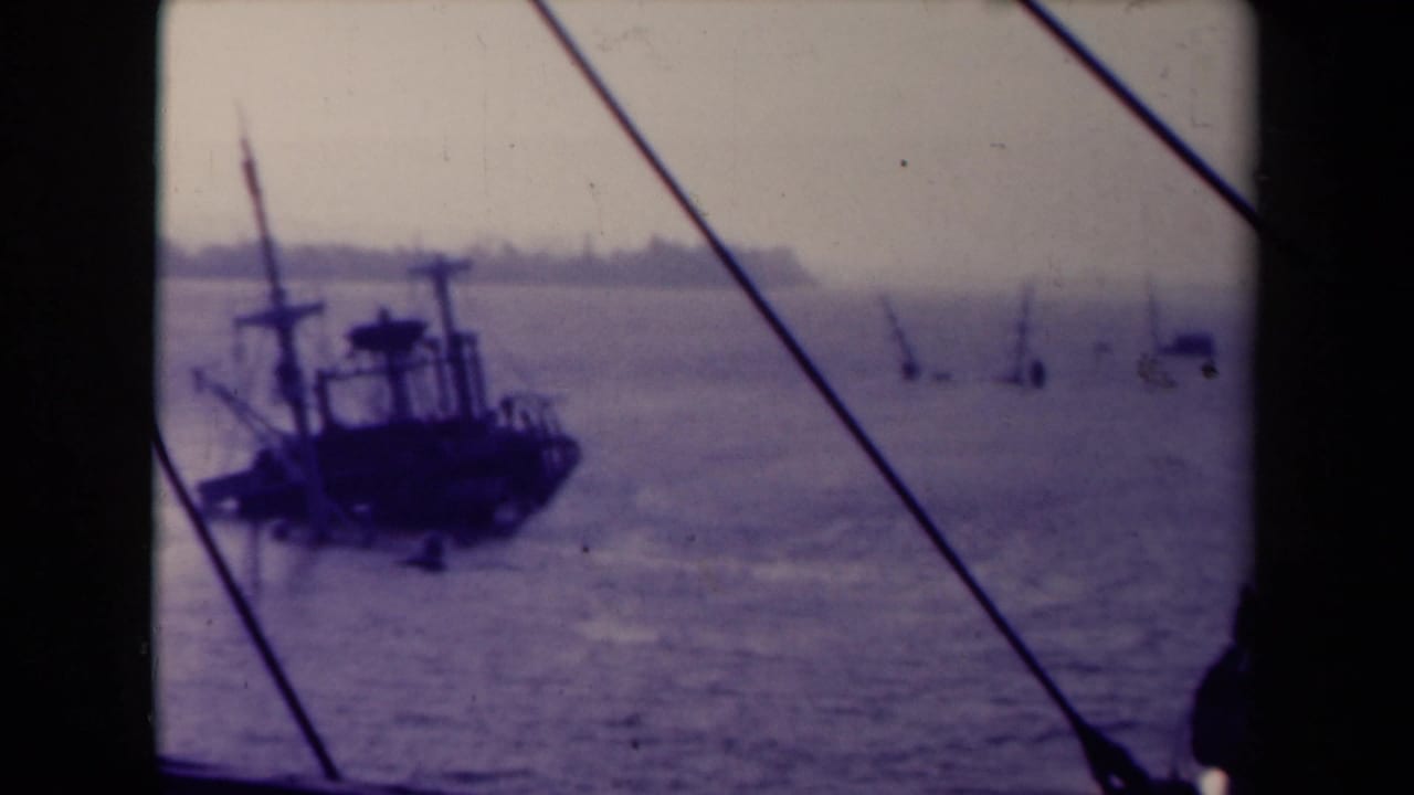 Dark working vessel and rigging seen across a hazy sea through a shipboard frame.
