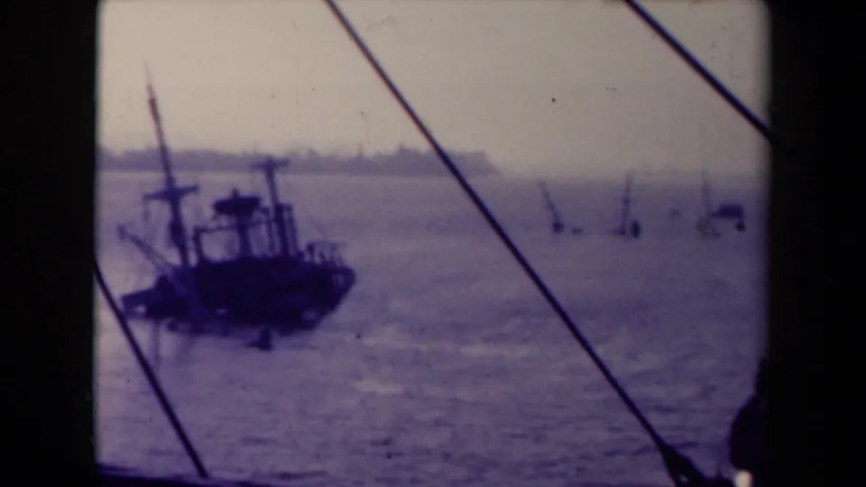 1945 France 8mm frame showing a dark vessel, diagonal rigging, distant shoreline, and hazy water from a moving maritime viewpoint.
