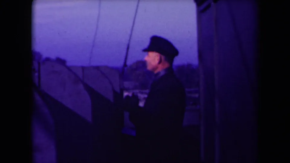 1945 France 8mm frame of an adult man in a cap, dark jacket and gloves standing at boat controls, seen from a nearby passenger angle.