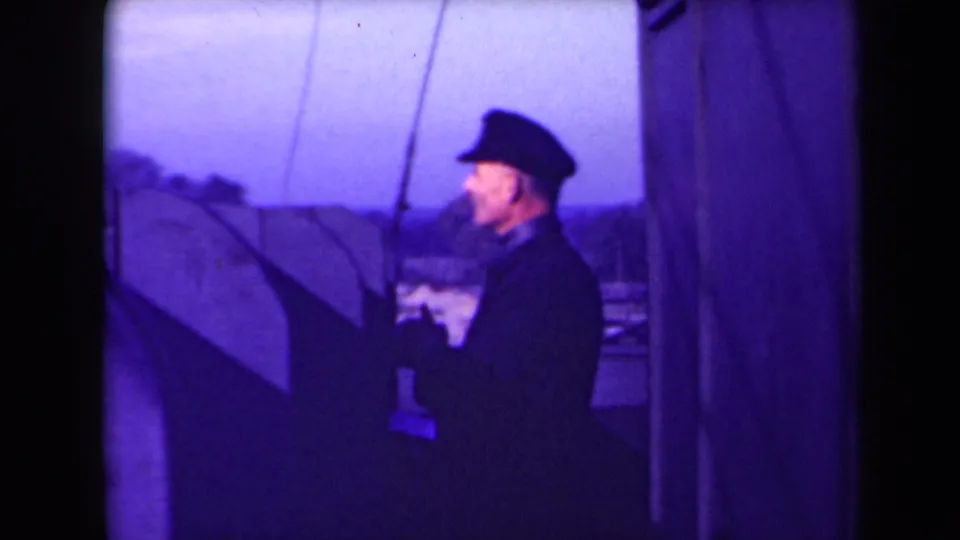 1945 France 8mm view of a man in cap, coat and gloves at a watercraft helm, with rigging and shoreline visible behind him.