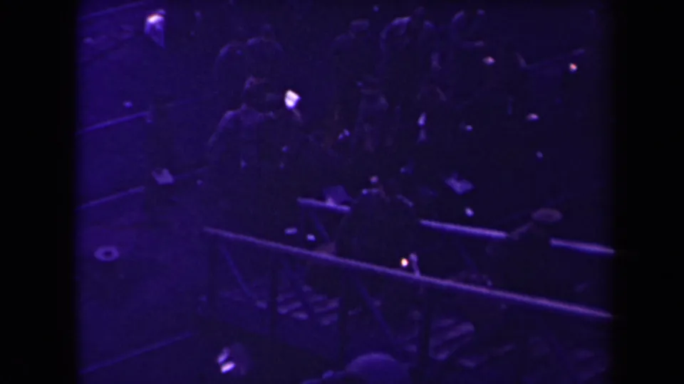 A low-light 8mm frame shows uniformed men with indistinct faces gathering by a dock or ship rail.