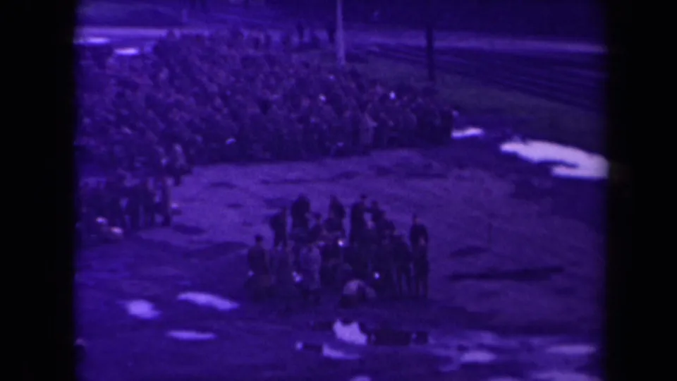 Wide nighttime view of uniformed soldiers gathered in rows on a dock or open quay, with rail lines or waterfront structures nearby.