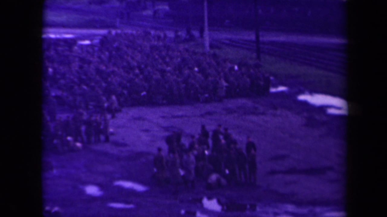 Distant crowd gathered on wet open ground with a smaller standing group in front.