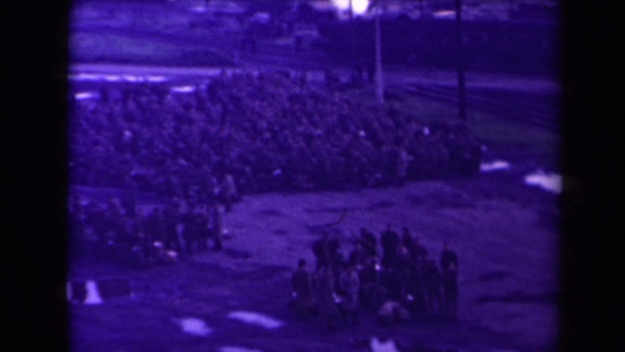 Large group sits in rows while a smaller cluster stands on the wet field.