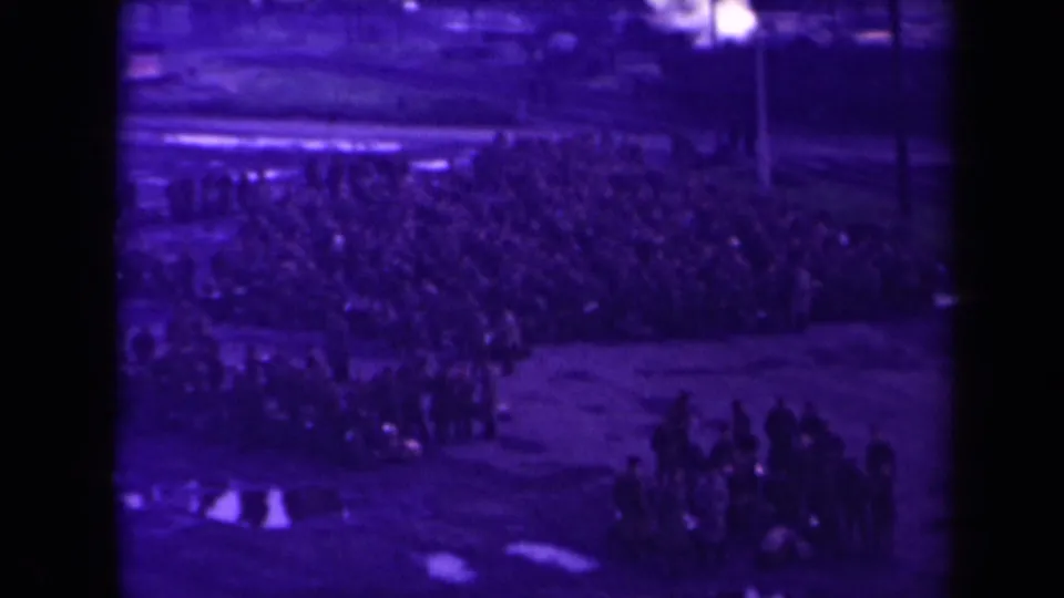Aged purple 8mm frame of a large outdoor assembly, with wet ground and distant figures in rows.