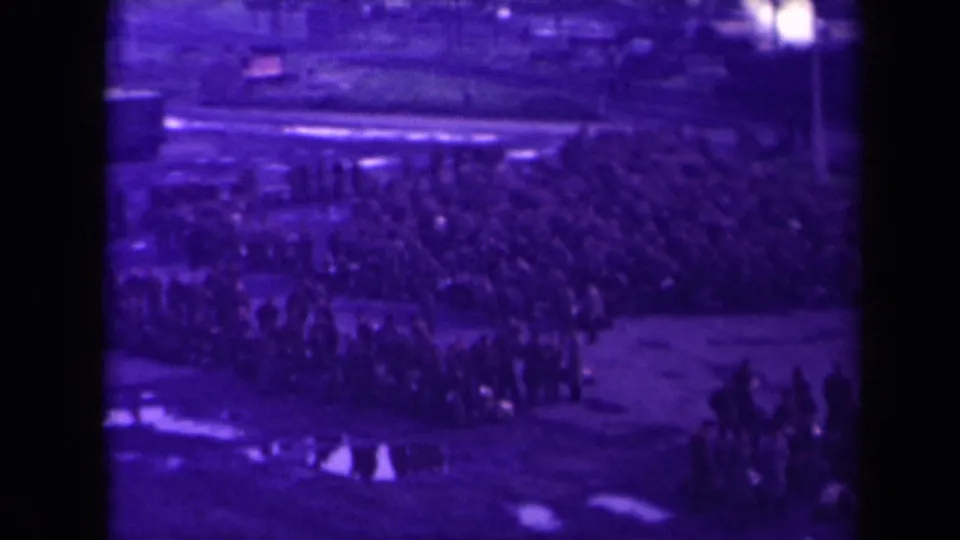 1945 France home-movie frame showing a mass outdoor gathering on muddy ground, heavily purple shifted by aged film.