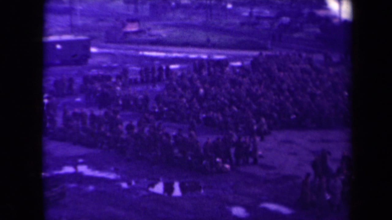 Large crowd gathered outdoors near rail-side ground with soldiers, civilians, luggage, and puddles.