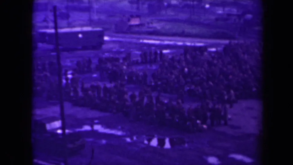 A dense line of people stands outdoors in 1945 France, with a rail car or station vehicle beyond the crowd and wet ground below.