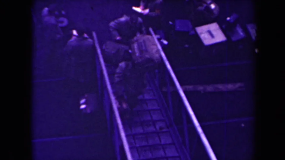 In 1945 France footage, soldiers appear on a metal ramp with railings inside a dark confined area.
