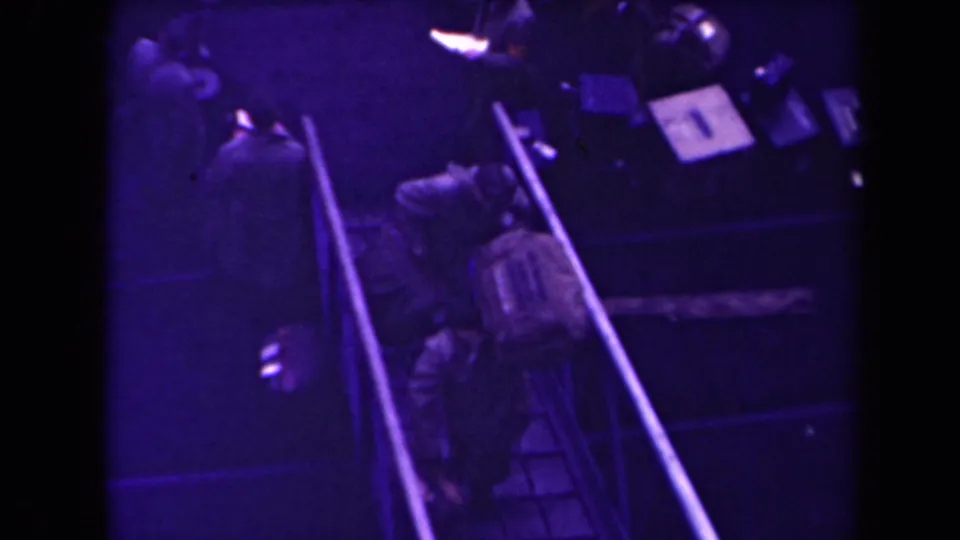 Soldiers in uniform carry or wear gear while moving through a narrow ramped passage in archival 1945 France footage.