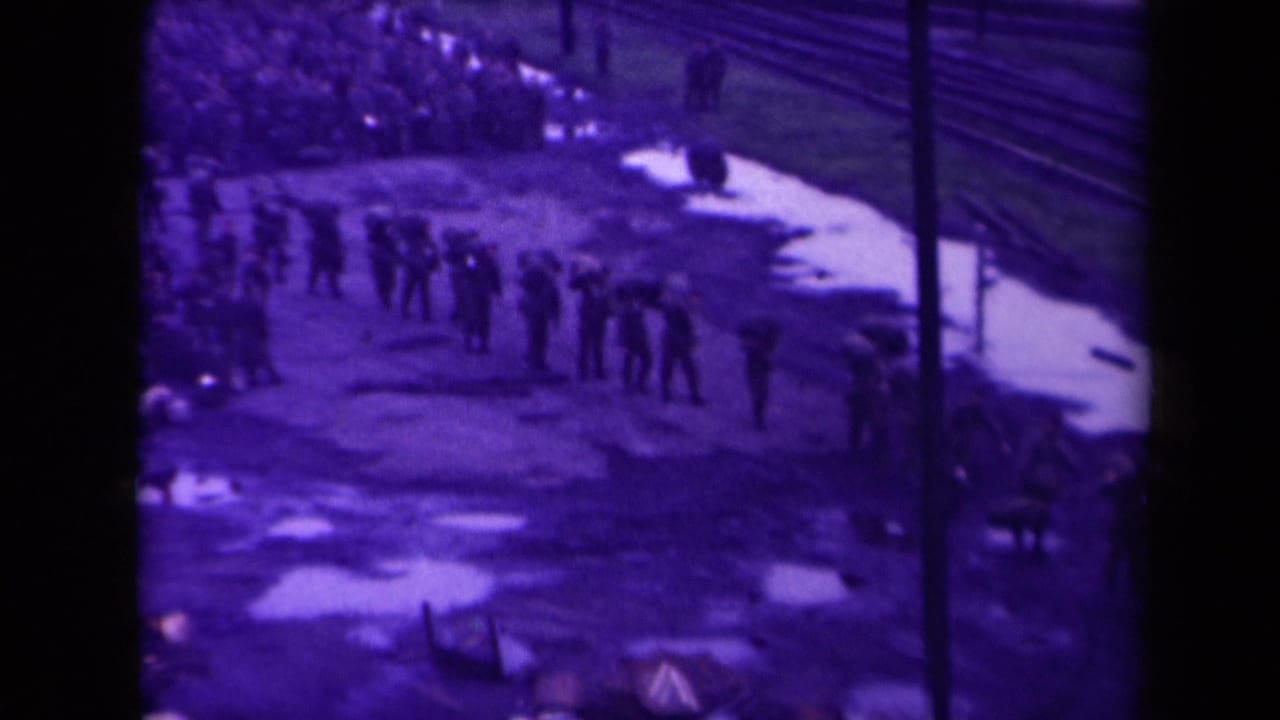 People stand and walk in loose lines beside railway tracks and pale wet ground.