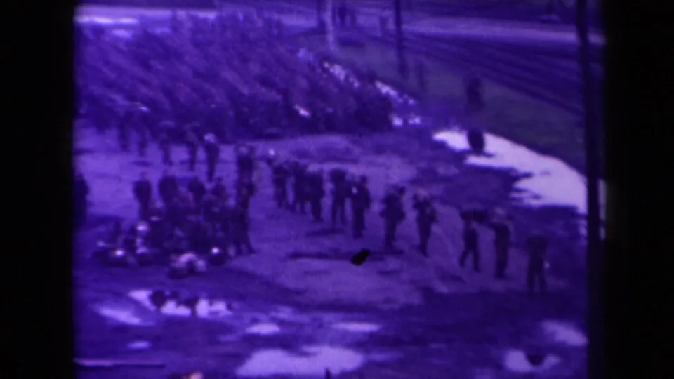 Purple-shifted 8mm frame of a large group near railway tracks, with individuals standing apart in a muddy French outdoor setting.