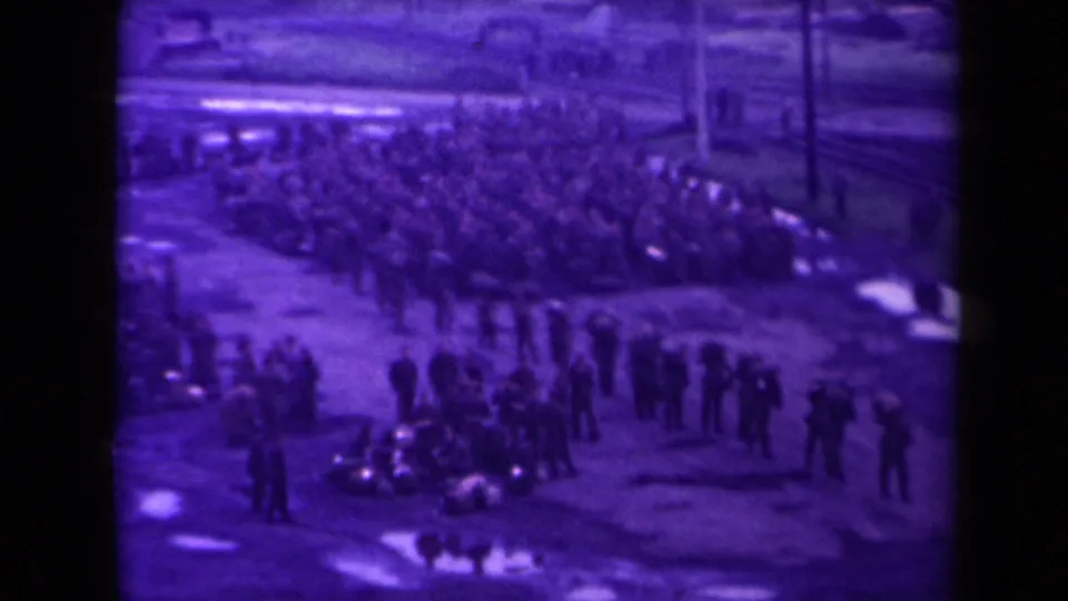Distant high-angle 1945 France home-movie frame showing a large crowd and smaller groups on muddy ground near rail lines.