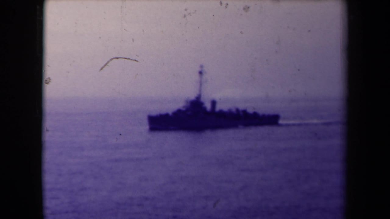 Dark naval ship crossing calm water beneath a hazy sky.