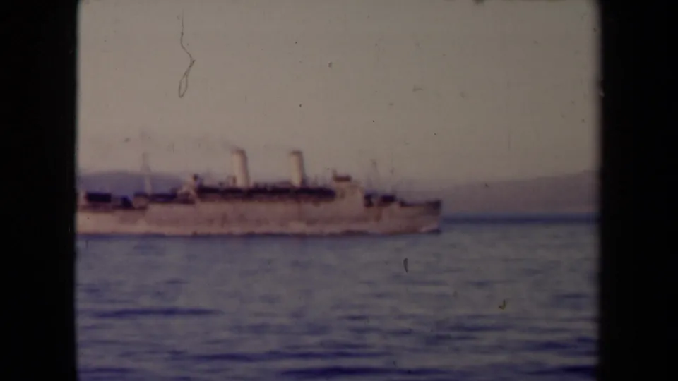 A larger light-colored transport or passenger vessel with several smokestacks passes across calm water, with hazy land in the distance.