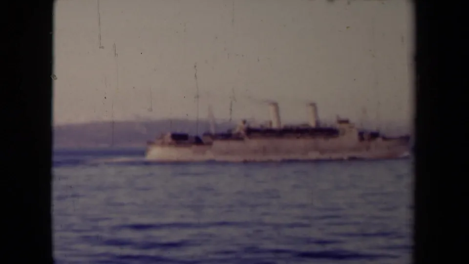 A soft 8mm view shows the steamship continuing across the water, with calm sea foreground and a low shoreline in the distance.