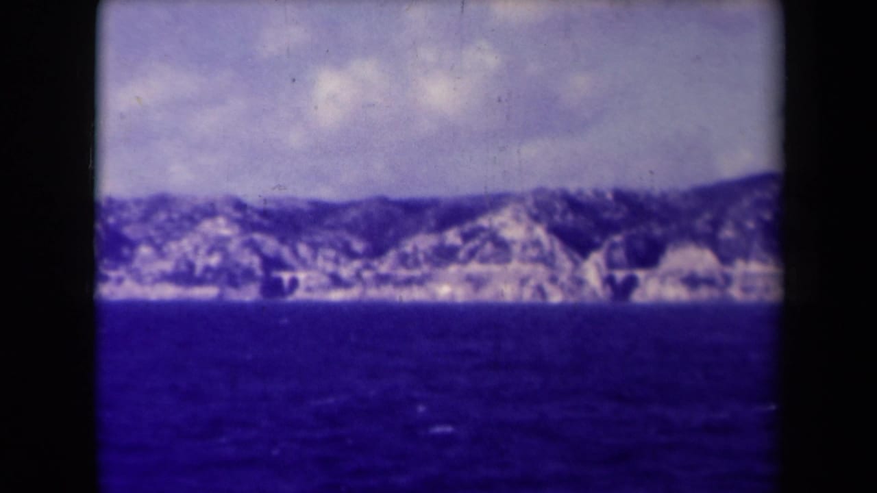 Rocky coastline and low mountains across blue water under clouds.