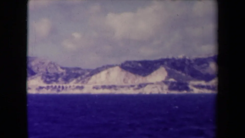 1945 France home-movie frame with cloudy sky, pale mountains, and dark blue water, seen from a moving amateur vantage point.