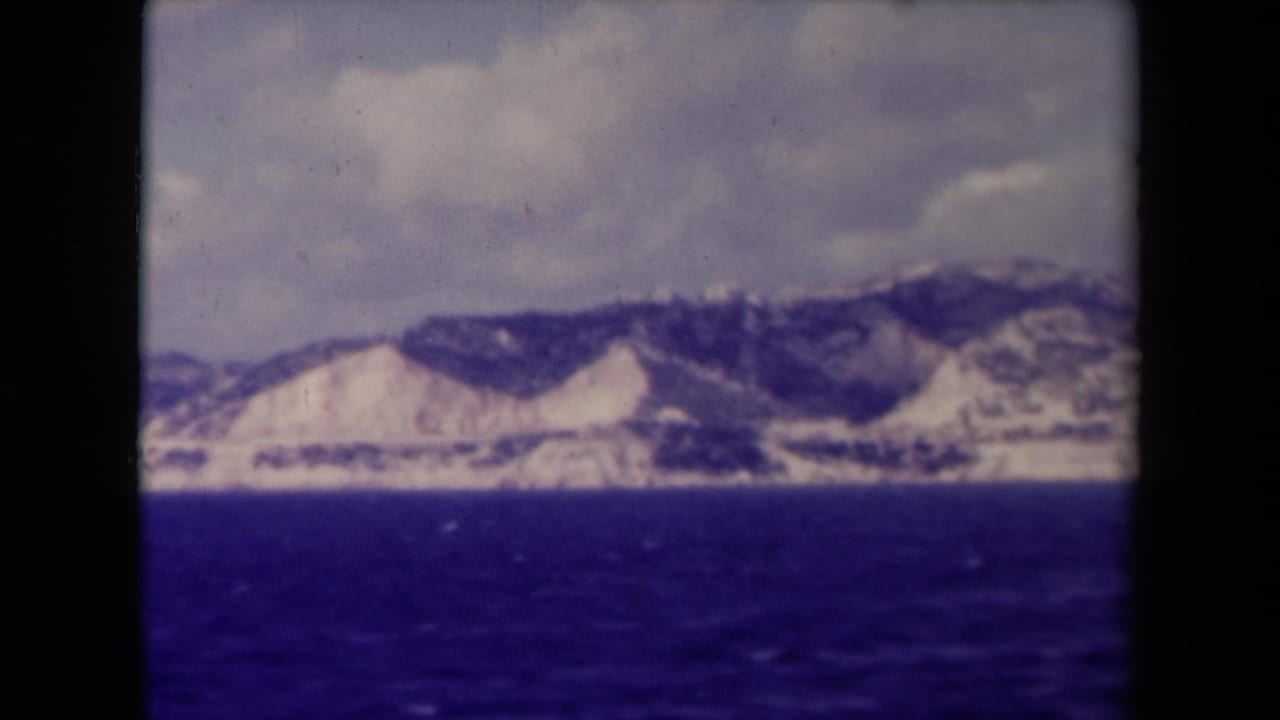 Distant rocky coastline and mountain ridge across a deep blue sea under clouds.