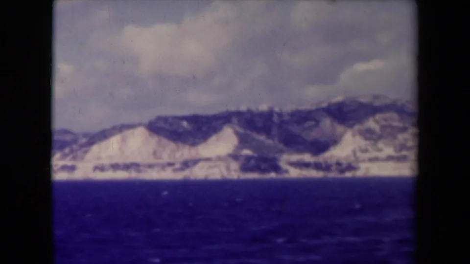 1945 8mm home-movie frame of a distant French coastal mountain range seen across open blue water beneath a cloudy sky.