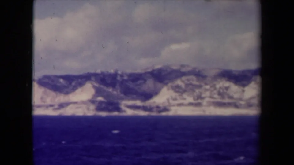 Soft 1945 8mm view of a rugged French shoreline from a vessel, with blue sea, pale hills, and clouded sky.