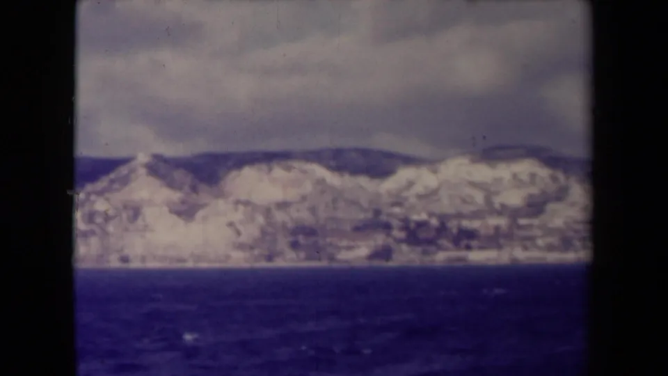 Final 1945 8mm frame of the French coast across the water, with light rocky slopes, darker hills, and cloud cover.