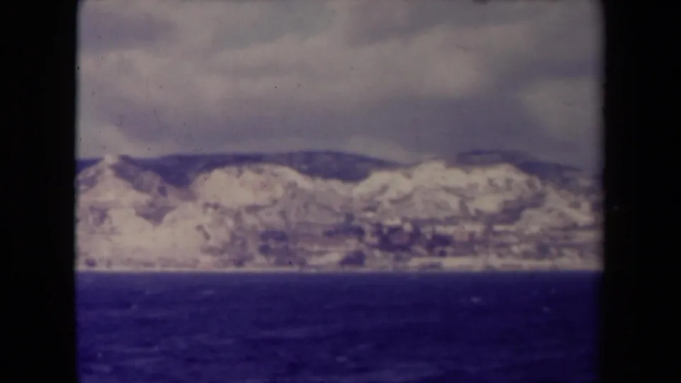 1945 France home-movie frame showing a distant snowy mountain range beyond dark blue water under overcast cloud.
