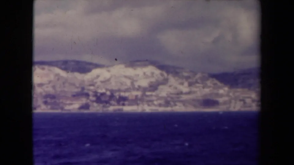 1945 France archival home-movie frame with a blurred snowy shoreline and mountains across deep blue water.