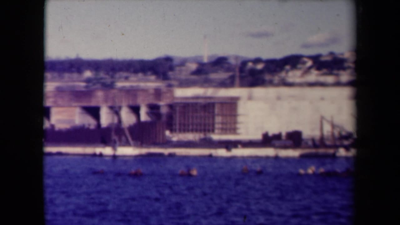 Waterfront construction site across blue water with a pale wall, cranes, small boats, and hillside buildings.