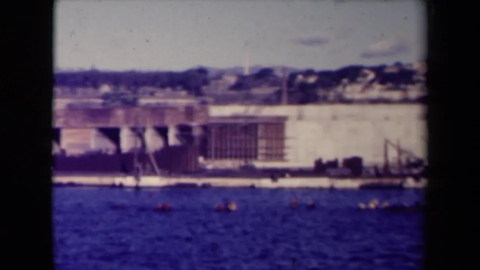 1945 France 8mm frame showing a waterfront worksite across blue water, with a pale wall, scaffolding, small boats, and hillside houses beyond.