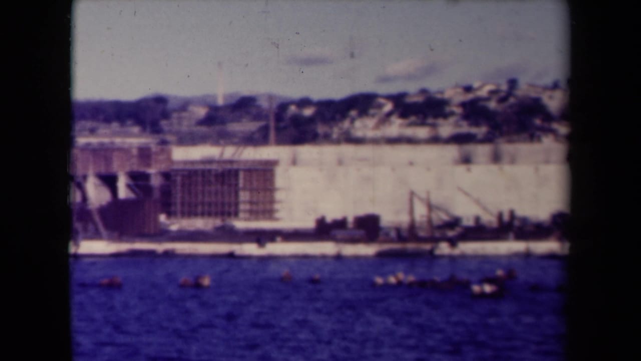 Shoreline wall and construction area with blue water, boats, cranes, scaffolding, and hillside buildings.