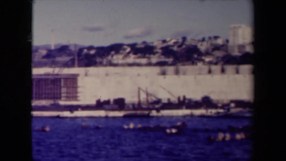 A 1945 France 8mm view across blue water toward a long waterfront wall, construction equipment, and hillside buildings under a bright sky.